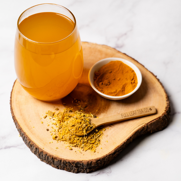 Glass of Organic Heartburn Relief Tea with vibrant golden color, wooden spoon of loose herbal blend, and bowl of turmeric on wooden slab with marble background.