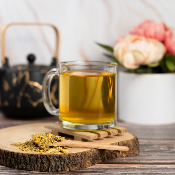 Glass cup of Organic Heartburn Relief Tea with golden herbal infusion, wooden spoon of loose leaf blend on wood slab, black teapot, and soft pink flowers in background.