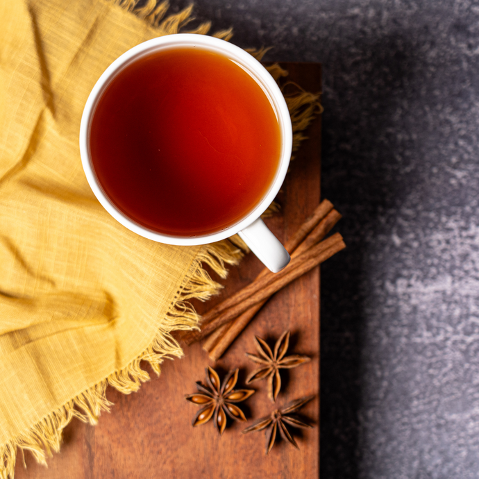 A cup of warm spiced tea on a wooden board with cinnamon sticks and star anise beside a yellow cloth, creating a cozy and inviting atmosphere.