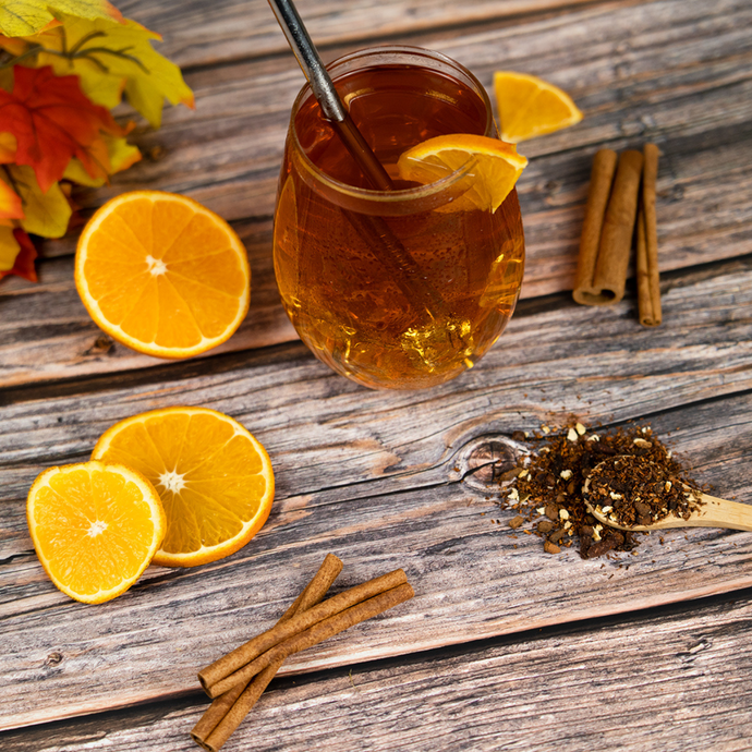Glass of iced tea garnished with an orange slice and metal straw, surrounded by orange slices, cinnamon sticks, and loose tea on a rustic wooden surface.