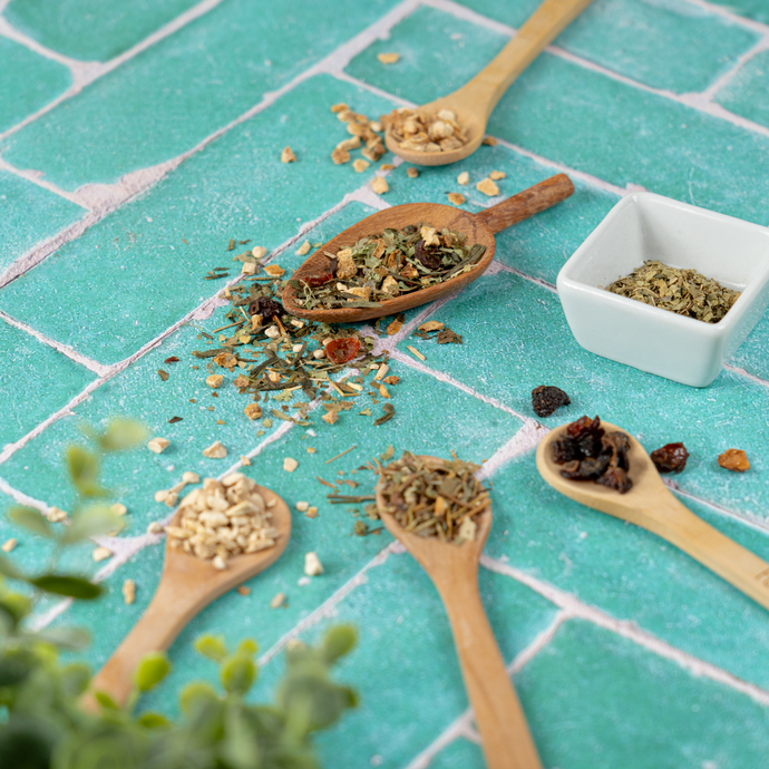 Assortment of loose Organic Live Fit Tea ingredients displayed in wooden spoons on a turquoise tile surface, featuring herbs, dried fruits, and roots.