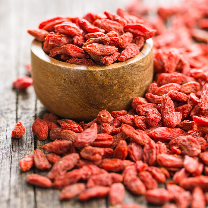 Close-up of vibrant red dried goji berries spilling from a wooden bowl onto a rustic wooden surface, symbolizing a nutrient-rich adaptogenic ingredient.