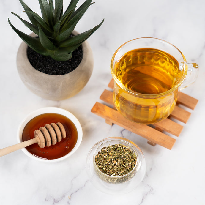Glass mug of tea with honey, dried herbs, and a small plant on a marble surface.