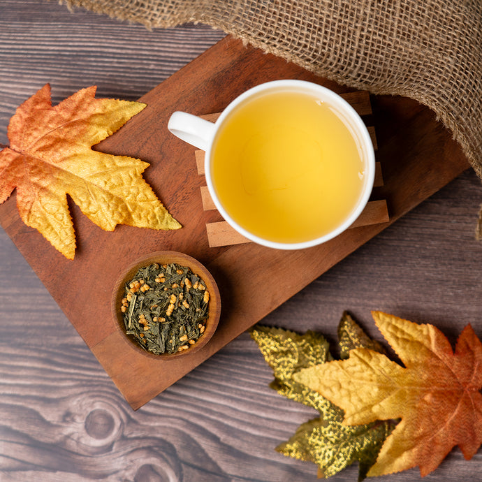 Tea cup with genmaicha tea, a small bowl of genmaicha tea leaves, and autumn leaves on a wooden board.