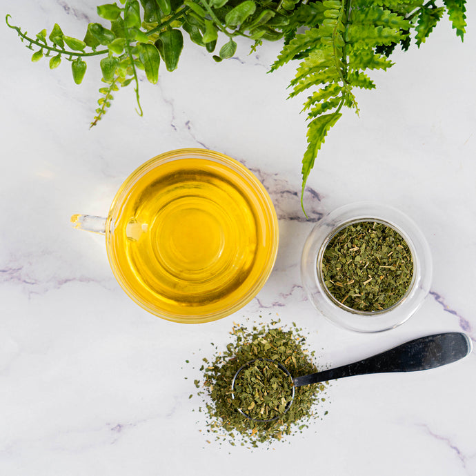 Tea cup with brewed tea, a spoonful of tea leaves, and a small container of dried tea leaves on a marble surface with ferns.