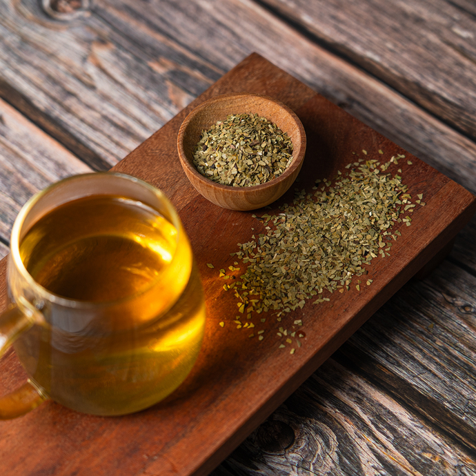 A glass mug of brewed Organic Yerba Mate tea on a wooden board with loose yerba mate leaves and a small wooden bowl of dried tea leaves beside it.