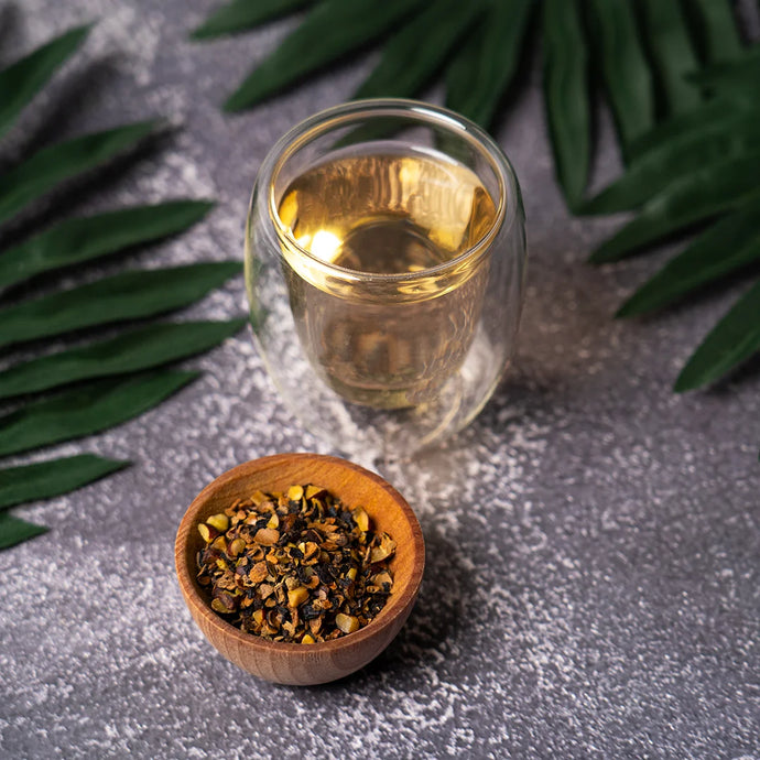 Glass of tea with a wooden bowl of tea leaves on a gray surface with green leaves.
