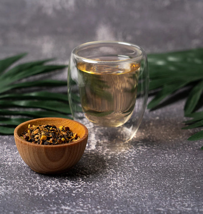 Clear glass cup of tea with a wooden bowl of tea leaves on a dark surface with green leaves in the background