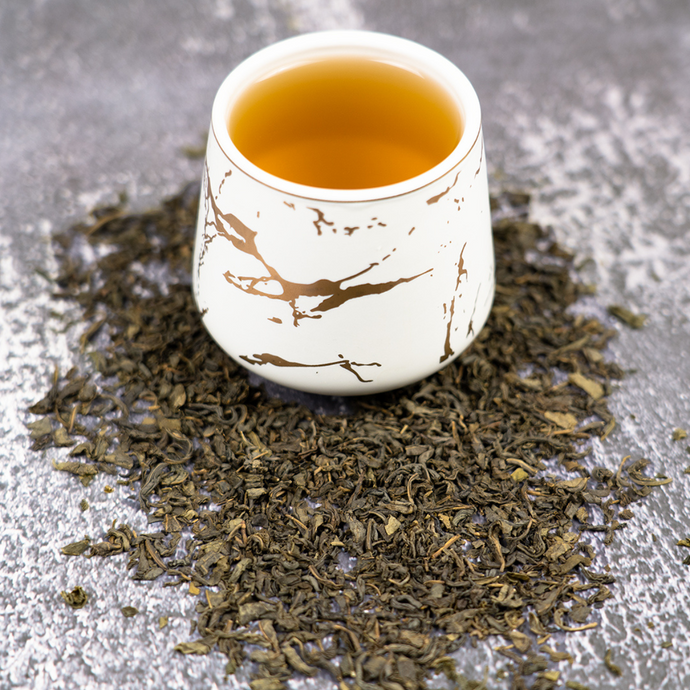 Cup of Organic Jasmine Tea in a white and gold marbled mug surrounded by loose jasmine green tea leaves on a textured gray surface.