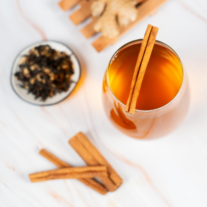A glass of Organic Masala Chai topped with cinnamon sticks, surrounded by loose-leaf tea, ginger pieces, and more cinnamon on a light marble surface.