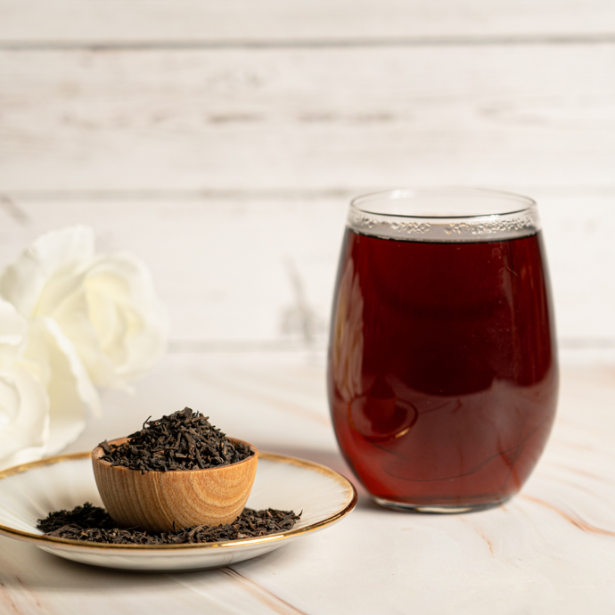 Glass of brewed Organic Pu-erh Tea with deep reddish-brown color beside a small wooden bowl filled with loose fermented tea leaves on a plate, set against a light wood background.
