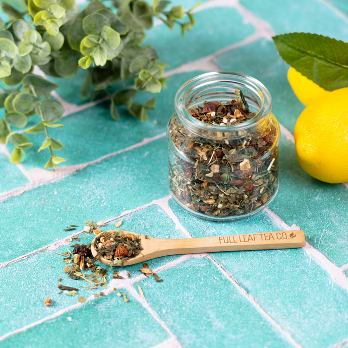 Glass jar and wooden spoon filled with loose Organic Live Fit Tea on a turquoise tile surface, surrounded by lemons and green leaves.