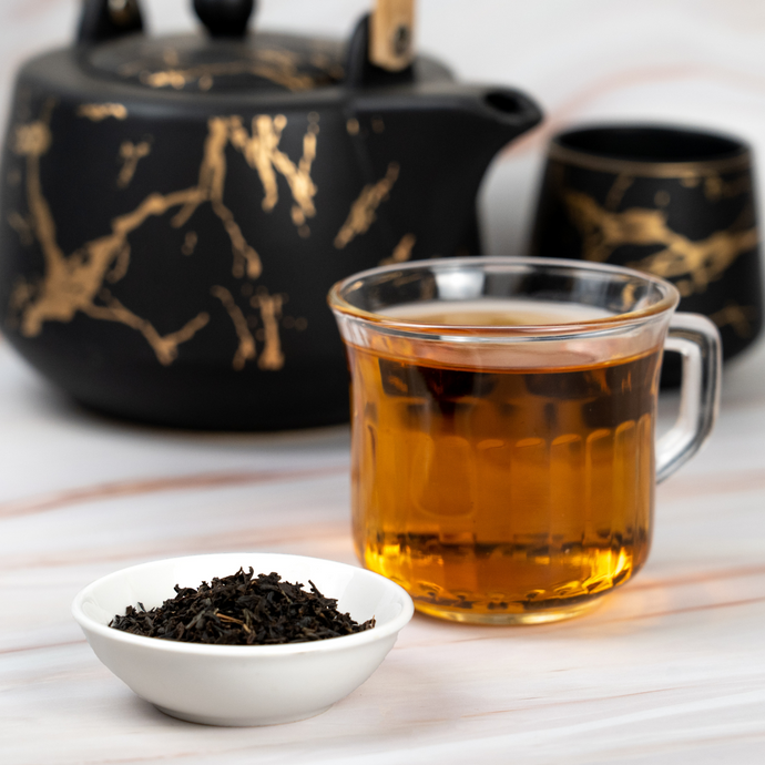 A glass cup of brewed black tea beside a small white bowl of loose-leaf tea, with an elegant black and gold teapot and matching cup in the background.