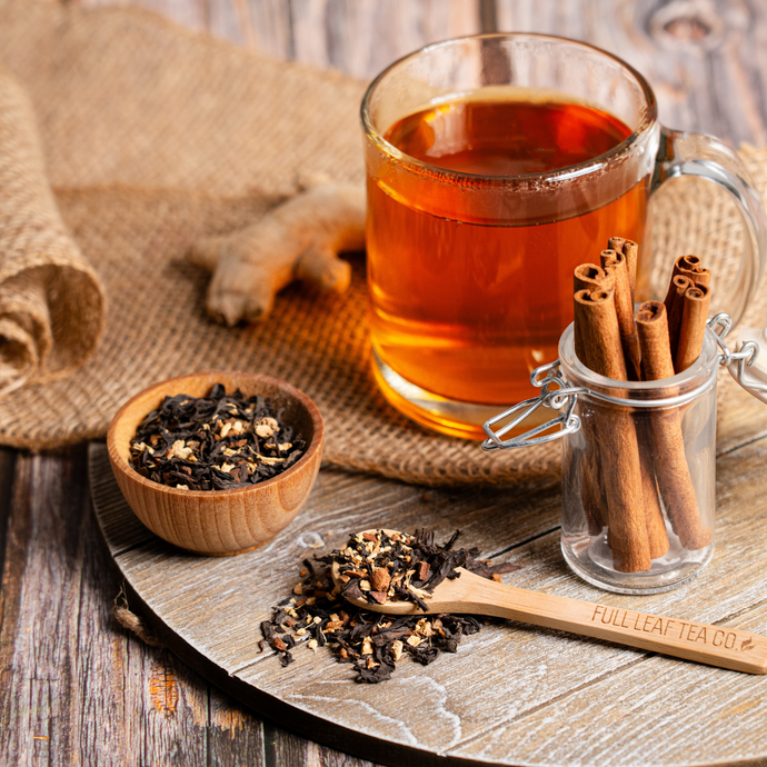 Glass mug of Organic Skinny Natural Tea with cinnamon sticks, loose leaf tea, and ginger on a rustic wooden surface.