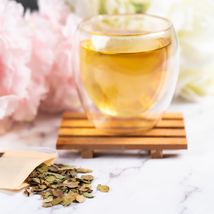 A clear double-walled glass cup of Organic Bilberry Leaf tea on a small wooden coaster, with a Full Leaf Tea Co. tea bag and dried bilberry leaves on a white marble surface, surrounded by soft pink and white flowers.