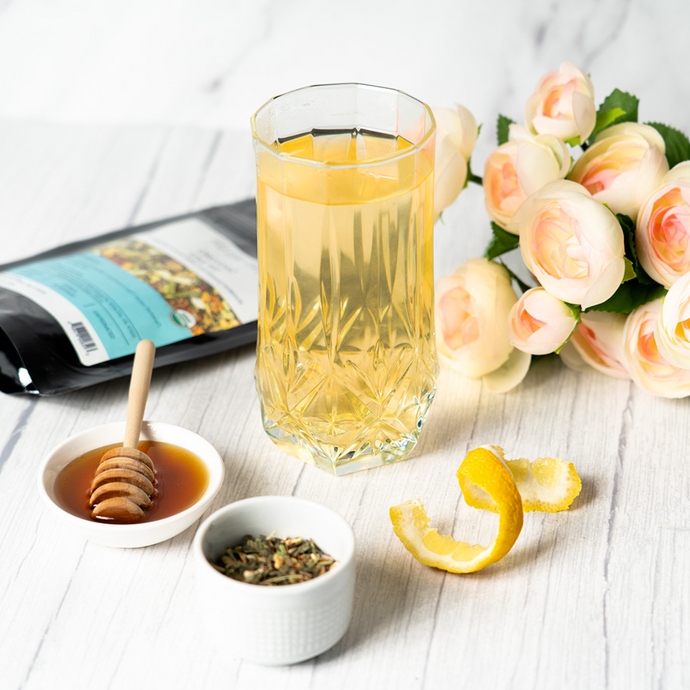 Glass of brewed Organic Live Fit Tea served with lemon peel, honey, and loose tea leaves, next to a floral arrangement and product pouch on a white wooden surface.