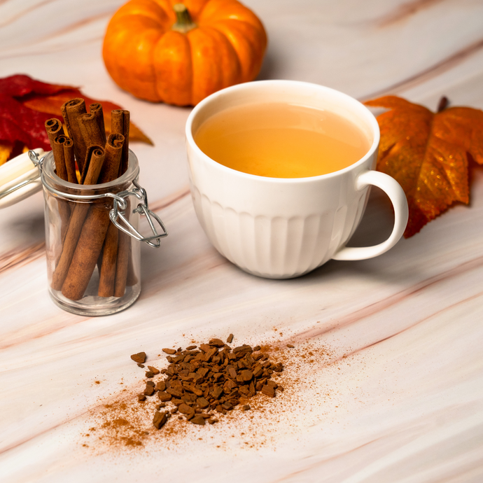 White cup of Organic Orange Cinnamon Spice tea beside a glass jar of cinnamon sticks, scattered spices, and small pumpkins with autumn leaves on a marble surface.