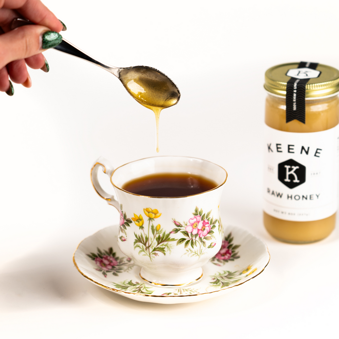 A cup of Organic English Breakfast Tea in a floral teacup as honey drips from a spoon, with a jar of Keene Raw Honey in the background.