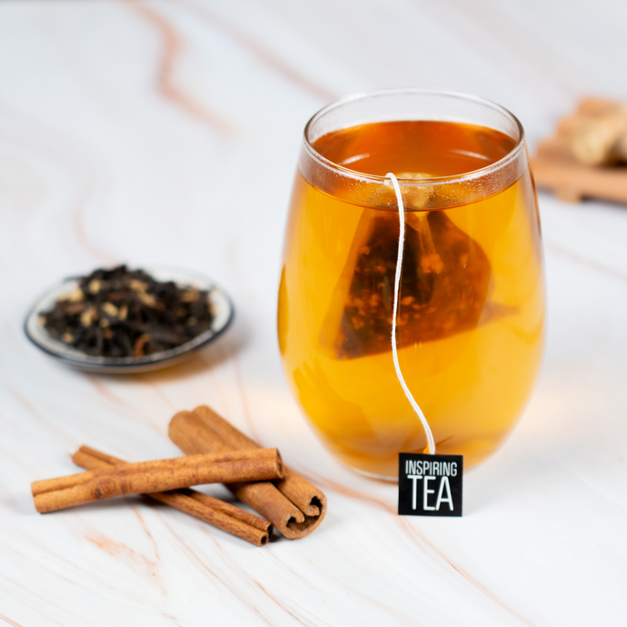 A glass cup of Organic Masala Chai with a tea bag steeping inside, surrounded by cinnamon sticks, loose-leaf spices, and ginger on a light marble surface.