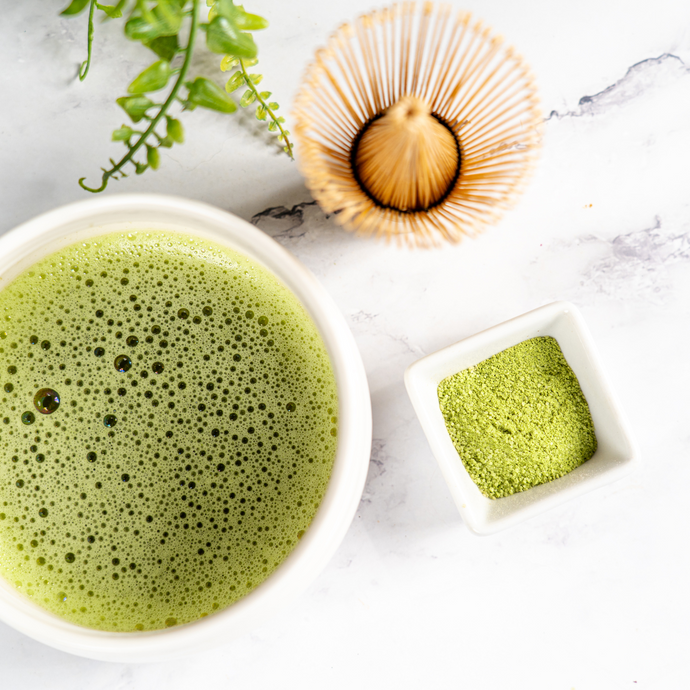 Whisked bowl of frothy green matcha tea with a bamboo whisk, small dish of vibrant matcha powder, and green leaves on a marble surface.