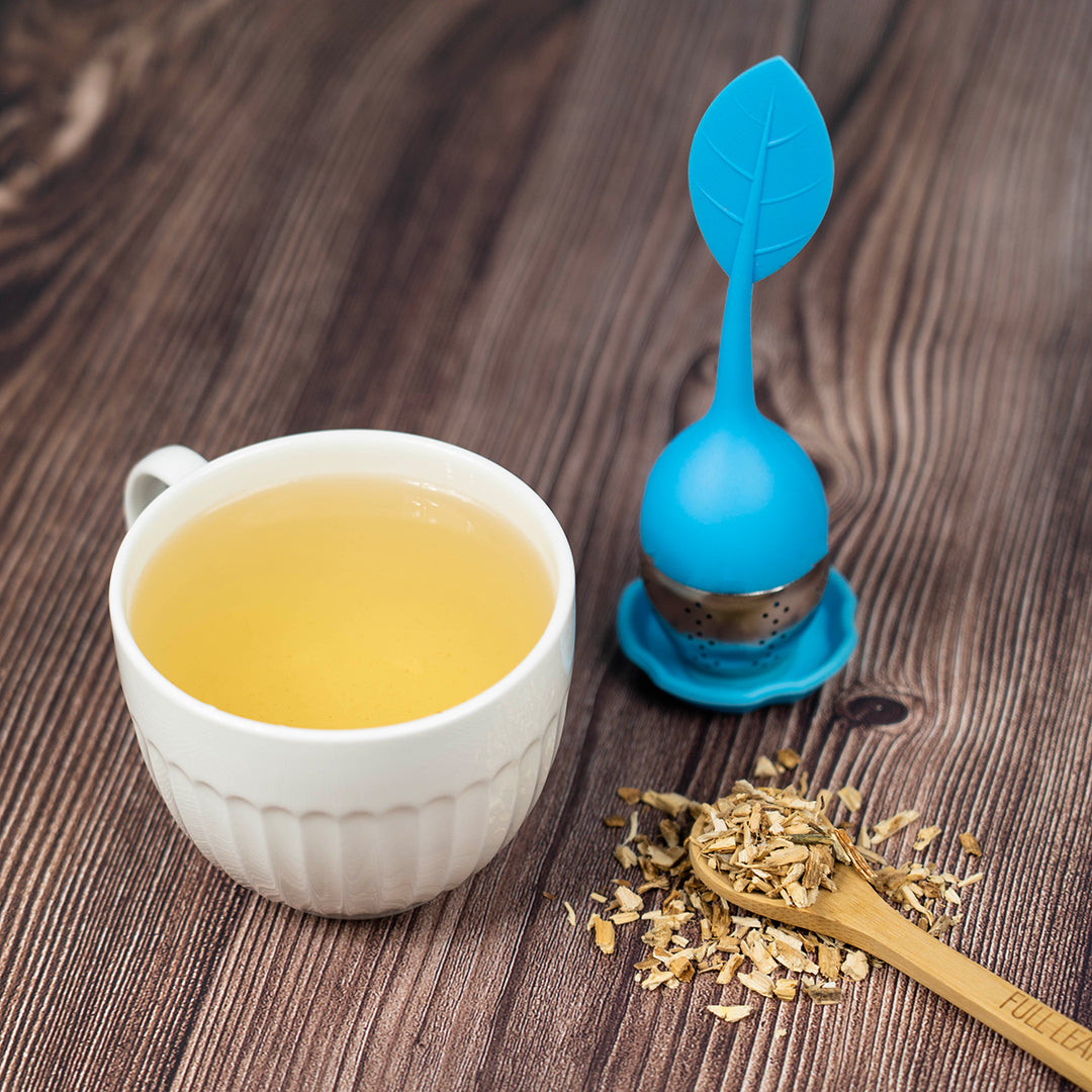 Tea cup with chicory root, a blue tea infuser, and a wooden spoon on a wooden surface.