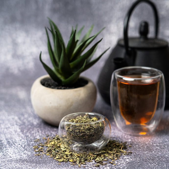 Tea leaves in a glass bowl with a cup of tea and a teapot on a gray surface.