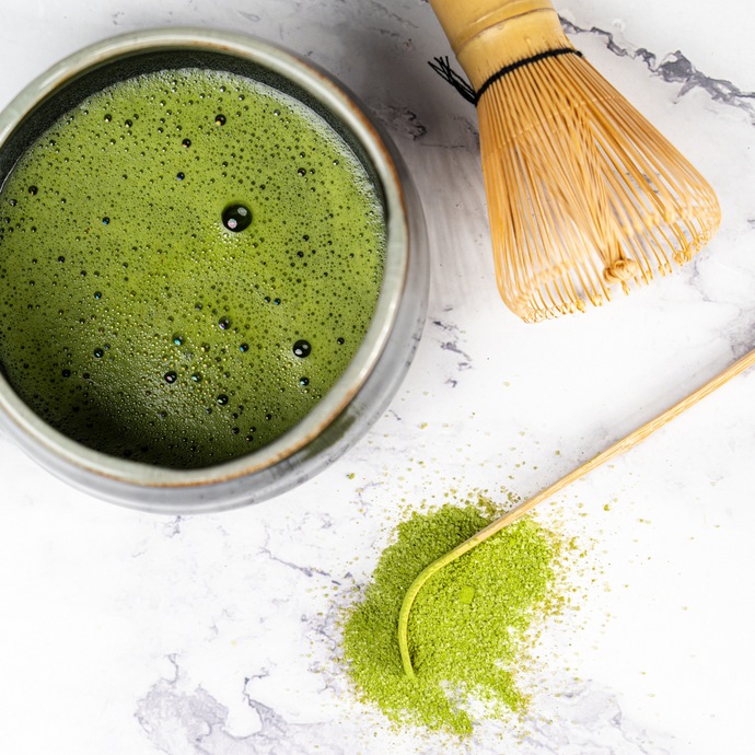 Traditional matcha tea preparation with a bowl of frothy green matcha, bamboo whisk, and bamboo scoop with vibrant matcha powder on a marble surface.
