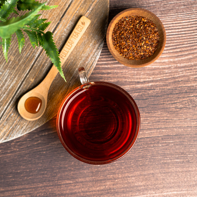 Cup of rooibos tea with a wooden spoon of honey and loose leaves on a wooden surface.
