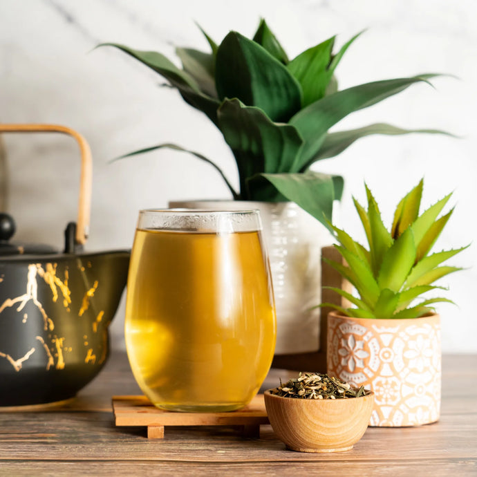 Glass of tea on a wooden table with a teapot, plants, and a small bowl of tea leaves.