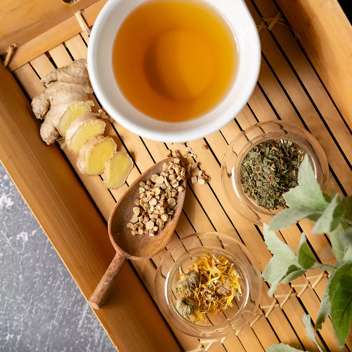 A white cup filled with lymphatic cleanse tea surrounded by ginger, and other ingredients in the blend on a wooden tray. 